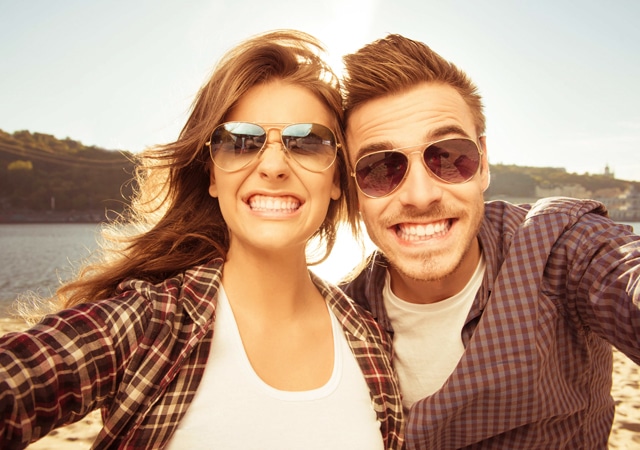 Two smiling people wearing sunglasses pose together outdoors by a lake on a sunny day.
