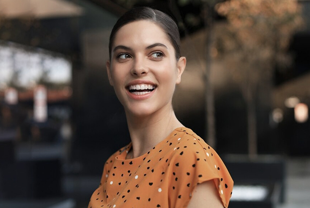 Smiling woman in orange polka dot shirt against blurred outdoor background.