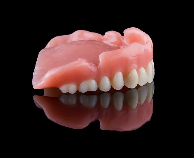 Denture prosthetic displaying artificial teeth and pink gum base against black background with reflection.