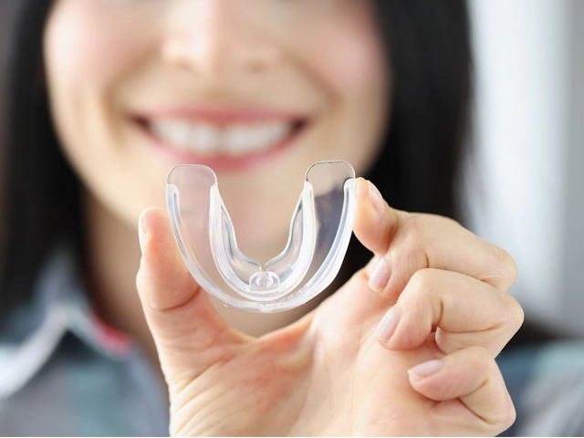 Woman smiling while holding clear dental aligner tray in clinical dental office setting.