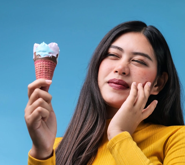 Young woman in yellow shirt holding ice cream cone against blue background, smiling