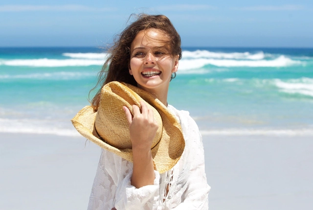 Woman holding hat smiling at beach in white shirt