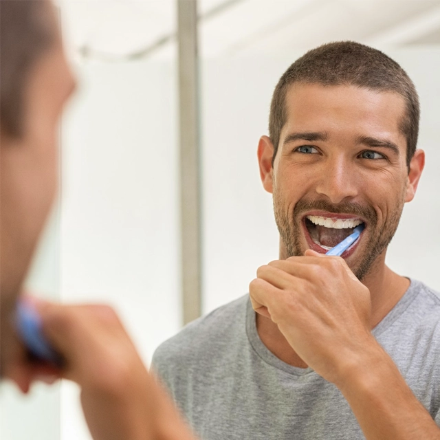 Man brushing teeth in bathroom mirror, smiling and practicing good dental hygiene.