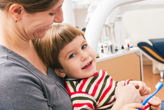 Adult holding smiling child in striped shirt at dental office.