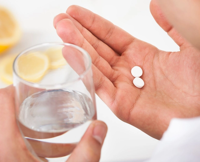 Hand holding two white pills above glass of water for medication consumption.