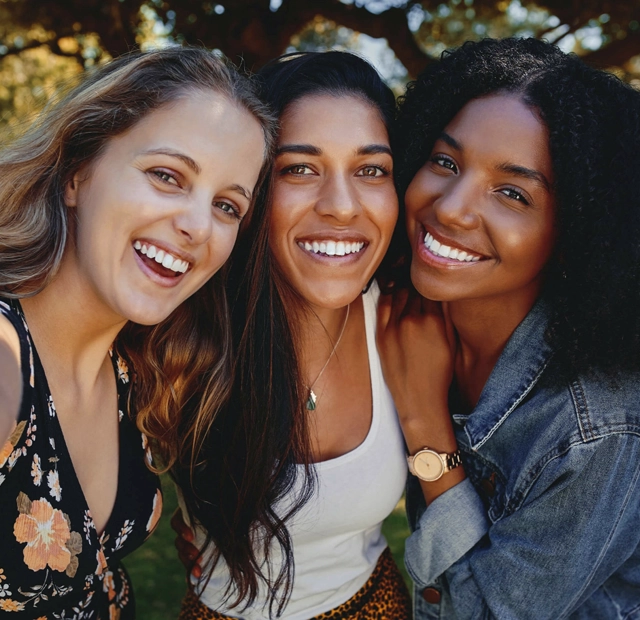 Three smiling women posing together outdoors in natural sunlight, showcasing healthy, bright smiles.