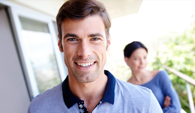 Smiling male dentist in blue shirt with colleague in background, professional dental office setting.