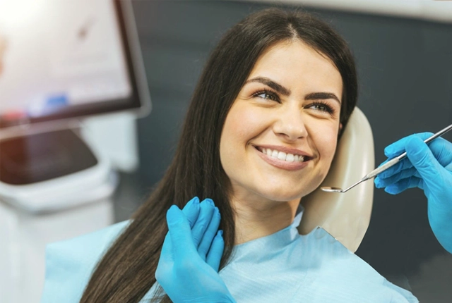 Female dental patient smiling in treatment chair wearing blue bib with dentist holding dental instruments.