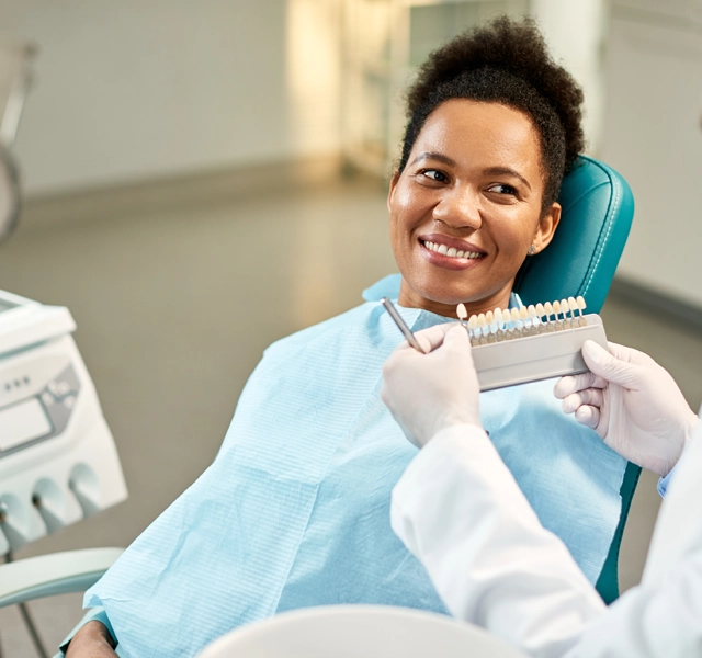 Smiling dental professional in blue bib holding shade guide in modern dental office.