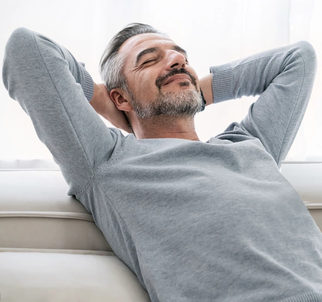 Relaxed middle-aged man reclined on couch with hands behind head, smiling contentedly indoors.