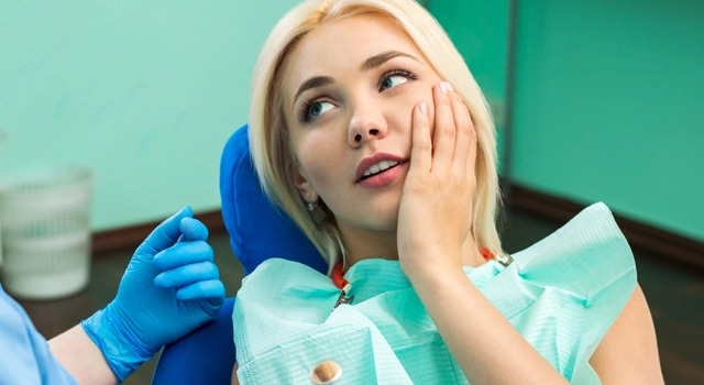 Woman in dental bib and blue gloves touching cheek in pain at dentist's office.