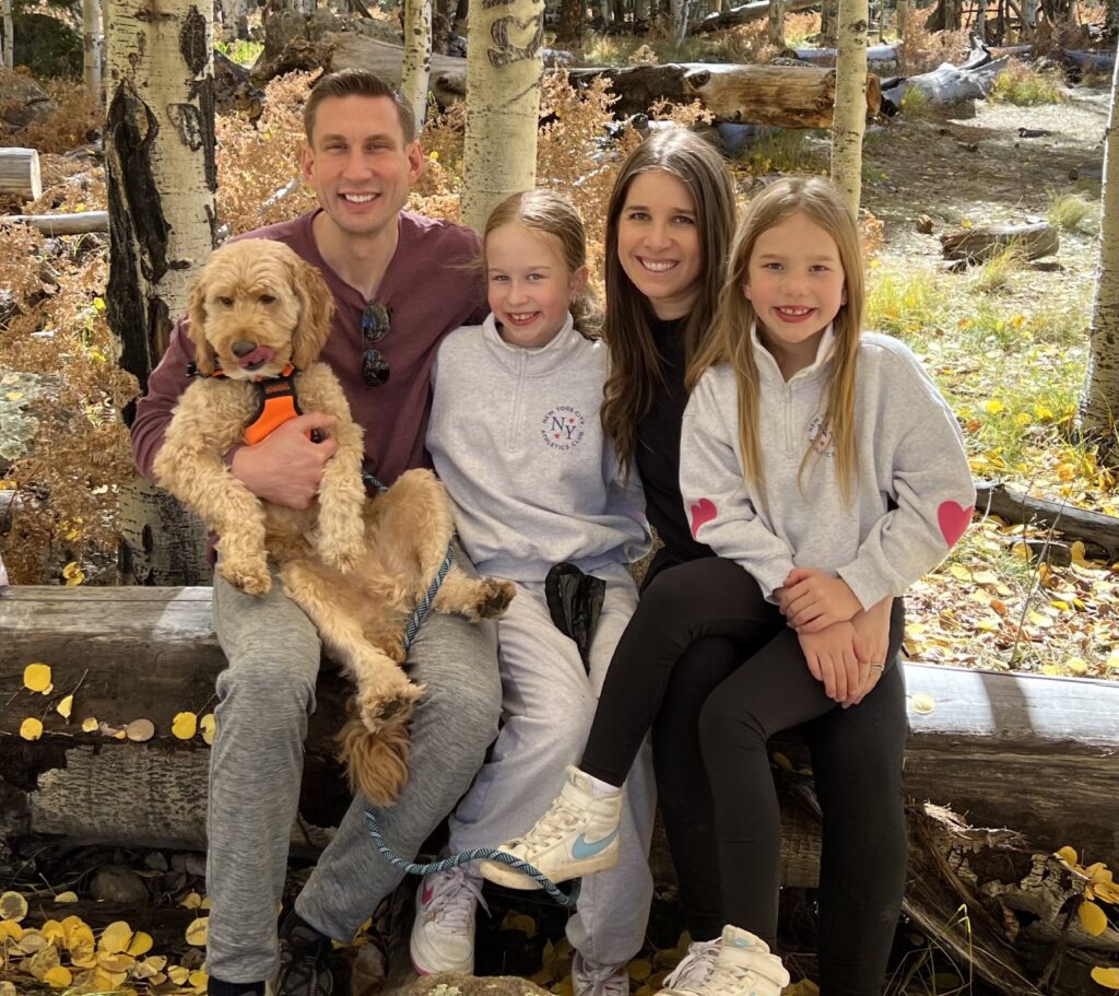 Family of five with dog posing together on outdoor wooden bench in fall setting.